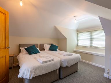 A bedroom with two single beds pushed together with white bedding teal pillows and folded towels by a window with blinds at Rocky Close in St Ives