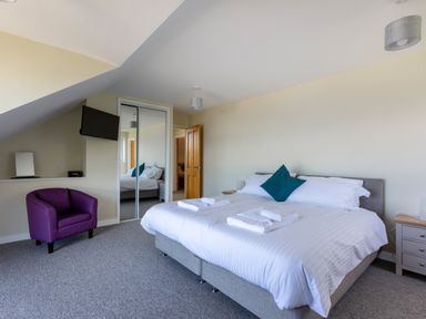 A bedroom with a double bed white linens and towels a purple armchair mounted TV and mirrored wardrobe at Rocky Close in St Ives