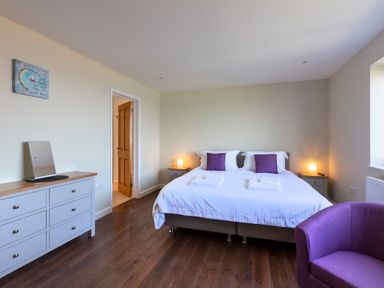 A bedroom with a double bed with purple pillows bedside tables with lamps a dresser with a mirror and a purple chair at Rocky Close in St Ives