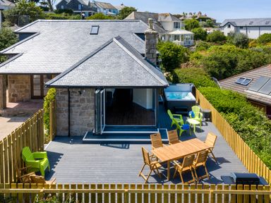 An outdoor deck with wooden dining table and chairs green plastic chairs a hot tub and barbecue at Rocky Close in St Ives