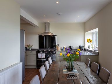 A kitchen with a wooden dining table with flowers on it and a black stove against the wall at Rocky Close in St Ives
