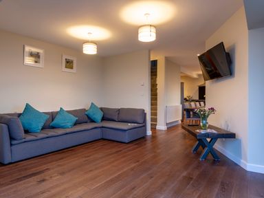 A living room with a gray sectional sofa with blue pillows two ceiling lights a wall-mounted TV and a wooden table with flowers at Rocky Close in St Ives