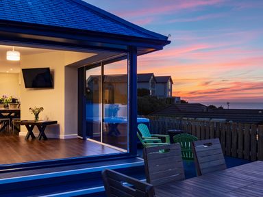 An open living and dining area with a wall-mounted television and wooden table inside a house with a wooden deck and outdoor chairs overlooking a sunset at Rocky Close in St Ives