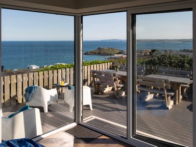 A view of a wooden deck with white chairs a small table and wooden picnic tables overlooking the sea through glass doors at Rocky Close in St Ives