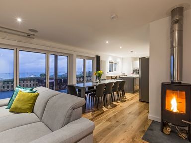 An open living space with a beige sofa with colorful pillows a dining table with chairs a wood stove and large windows overlooking a patio at Chy Lowen in St Ives