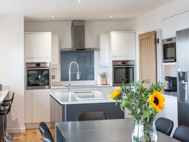 A modern kitchen with white cabinets, two built-in ovens, a kitchen island with a faucet, and a dining table with a vase of sunflowers at Chy Lowen in St Ives