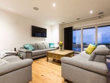 A living room with three gray sofas a wooden coffee table a wall-mounted TV and sliding glass doors at Chy Lowen in St Ives