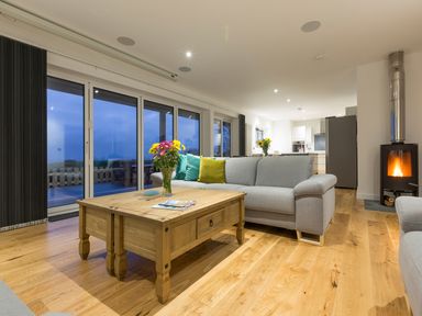 A living room with a wooden coffee table and grey sofas with pillows near a fireplace and large glass doors at Chy Lowen in St Ives