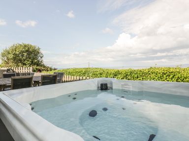 A hot tub filled with water on a patio next to a wooden dining table and chairs with greenery and sky in the background at Chy Lowen in St Ives