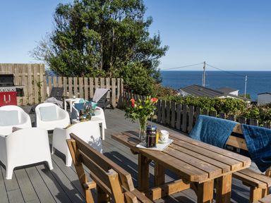 An outdoor deck with wooden table and benches white chairs and loungers overlooking the sea at Chy Lowen in St Ives