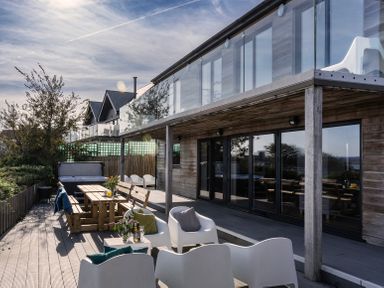 A patio with wooden tables and white chairs outside a modern house with glass balcony at Chy Lowen in St Ives