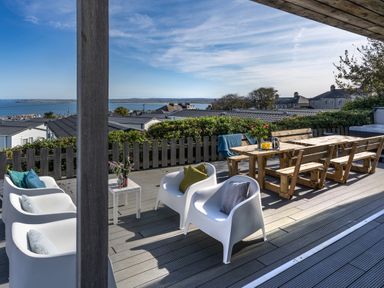 An outdoor deck with white chairs, a wooden dining table with benches, plants and ocean view at Chy Lowen in St Ives
