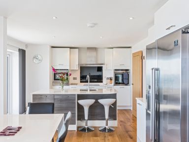 A modern kitchen with a kitchen island, two white bar stools, stainless steel refrigerator, built-in ovens, and a dining table at Boscarne in St Ives