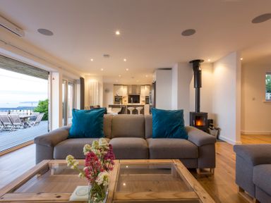 A living room with a gray sofa with blue pillows and a glass coffee table with flowers and an open view to a kitchen and dining area at Boscarne in St Ives