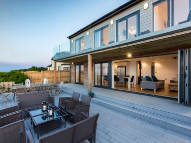 An outdoor deck with seating and tables connected to a two-story house with large glass doors open at Boscarne in St Ives