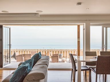 A living room with a sofa and dining table opening to an outdoor deck with seating and ocean view at Boscarne in St Ives