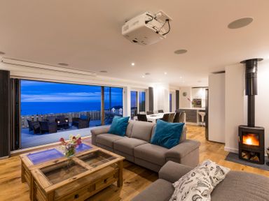 A living room with sofas and blue cushions near a fireplace and glass doors opening to an outdoor seating area with ocean views at Boscarne in St Ives