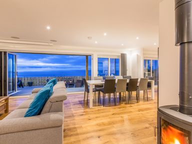 An open living and dining area with a beige sofa and blue cushions a dining table with eight chairs wooden flooring and a wood-burning stove next to large folding doors opening to a terrace with ocean view at Boscarne in St Ives