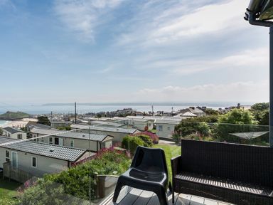 An outdoor balcony with black plastic chair and wicker loveseat overlooking a caravan park and coastal town at Boscarne in St Ives
