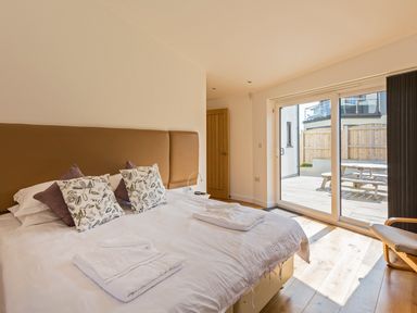 A bedroom with a large bed with pillows and folded towels near a sliding glass door leading to an outdoor area with a wooden table at Boscarne in St Ives