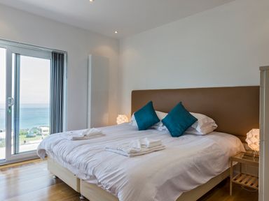 A bedroom with a double bed with white bedding and blue pillows near a sliding glass door with ocean view at Boscarne in St Ives