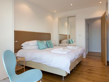 A bedroom with a double bed with white linens and teal pillows a blue chair and mirrored wardrobe at Boscarne in St Ives