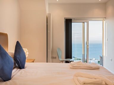 A bedroom with a bed with blue pillows towels on it and a chair near a sliding glass door with ocean view at Boscarne in St Ives