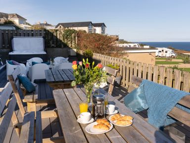 An outdoor wooden patio with tables and benches croissants and drinks on table a hot tub and chairs in the background and ocean view at Boscarne St Ives