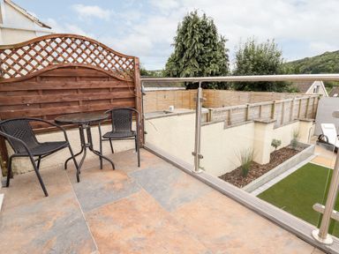 A balcony with a round table and two chairs overlooking a fenced garden with trees at 4 Bodnant Road in Rhos-On-Sea