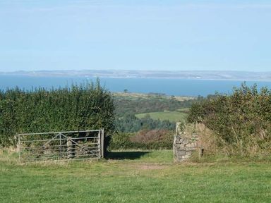 A metal gate and stone post opening to a grassy field with hedges and a view of the sea in the distance at The Dovecote in Hartland