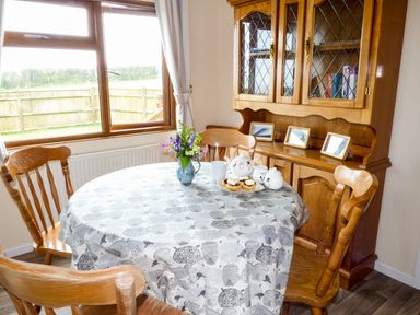 A dining room with a round table set with a tablecloth and tea set surrounded by wooden chairs and a wooden cabinet with framed pictures at The Dovecote in Hartland