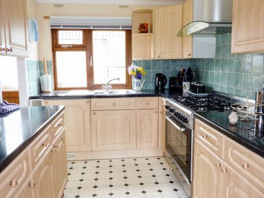 A kitchen with wooden cabinets a gas stove a sink under a window and green tiled backsplash at The Dovecote in Hartland