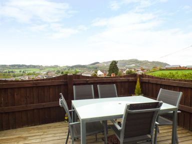 An outdoor patio area with a glass table and six chairs surrounded by a wooden fence with a view of hills and houses at Penylodge in Builth Wells
