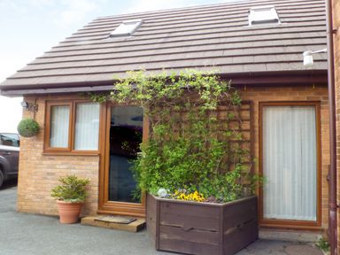 The exterior of a brick house with wooden framed windows and door with a large wooden planter with flowers and climbing plants at Penylodge in Builth Wells