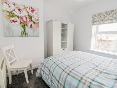 A bedroom with a bed covered in a plaid duvet a white chair a white wardrobe a window with patterned blind and floral painting on the wall at Sunnie Cottage in Seahouses