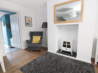 A sitting area with a gray armchair holding a yellow pillow a floor lamp a white stove in a recessed wall space a large rectangular mirror above the stove and a black and white rug on a wooden floor at Sunnie Cottage in Seahouses