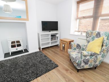 A living room with a patterned armchair a yellow cushion a television on a white stand a small stack of nested wooden tables a rug and an electric fireplace at Sunnie Cottage in Seahouses