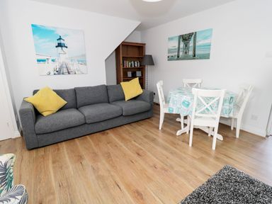A living area with a grey sofa with yellow cushions a wooden floor a round dining table with four chairs and two wall pictures at Sunnie Cottage in Seahouses