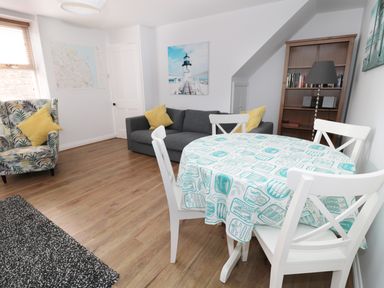 A living room with a round dining table covered with a blue and white tablecloth four white chairs a grey sofa with yellow pillows a patterned armchair a wooden bookshelf and a floor lamp at Sunnie Cottage in Seahouses