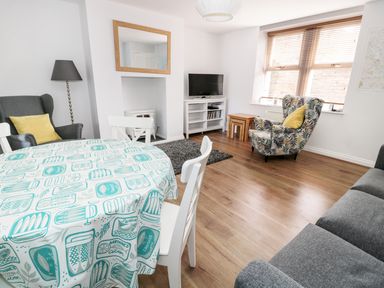 A living room with a dining table covered with a patterned tablecloth white chairs a grey armchair with a yellow cushion a patterned armchair with a yellow cushion a television on a white stand wooden flooring and a window with wooden blinds at Sunnie Cottage in Seahouses