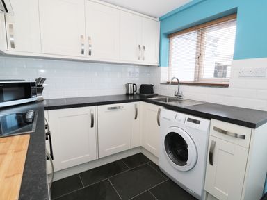 A kitchen with white cabinets black countertops a washing machine microwave kettle toaster and a window with blinds at Sunnie Cottage in Seahouses