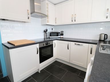A kitchen with white cabinets black countertops an oven microwave knife block kettle and a cutting board at Sunnie Cottage in Seahouses
