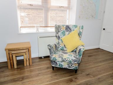 A patterned armchair with a yellow cushion next to nesting wooden tables under a window at Sunnie Cottage in Seahouses