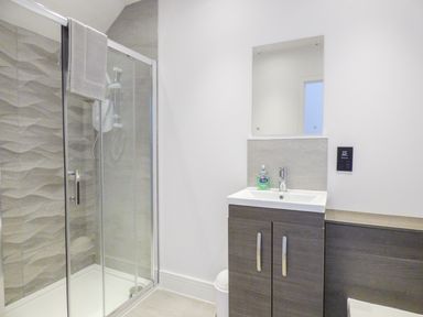 A bathroom with a glass shower enclosure a towel hanging on the shower door a vanity with a sink and mirror a soap dispenser and a window at 5 Ocean Heights in Newquay