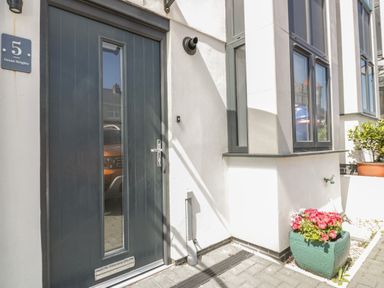 A black front door with a glass panel next to a white wall and windows with a green planter holding pink and white flowers at 5 Ocean Heights in Newquay