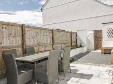 An outdoor patio with a glass top table and six wicker chairs next to a wooden fence at 5 Ocean Heights in Newquay