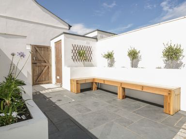 An outdoor patio with wooden bench seating plants in raised white planters and wooden doors at 5 Ocean Heights in Newquay