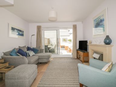 A living room with a grey sofa and ottoman a blue armchair television fireplace and sliding glass doors leading outside at 5 Ocean Heights in Newquay