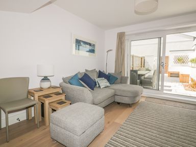 A living room with a gray sofa with pillows a small gray ottoman nested wooden tables a lamp a tan chair a floor lamp and sliding glass doors leading to a patio at 5 Ocean Heights in Newquay