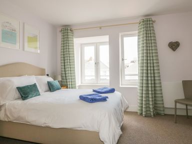 A bedroom with a bed with white sheets and blue towels near windows with patterned curtains at 5 Ocean Heights in Newquay